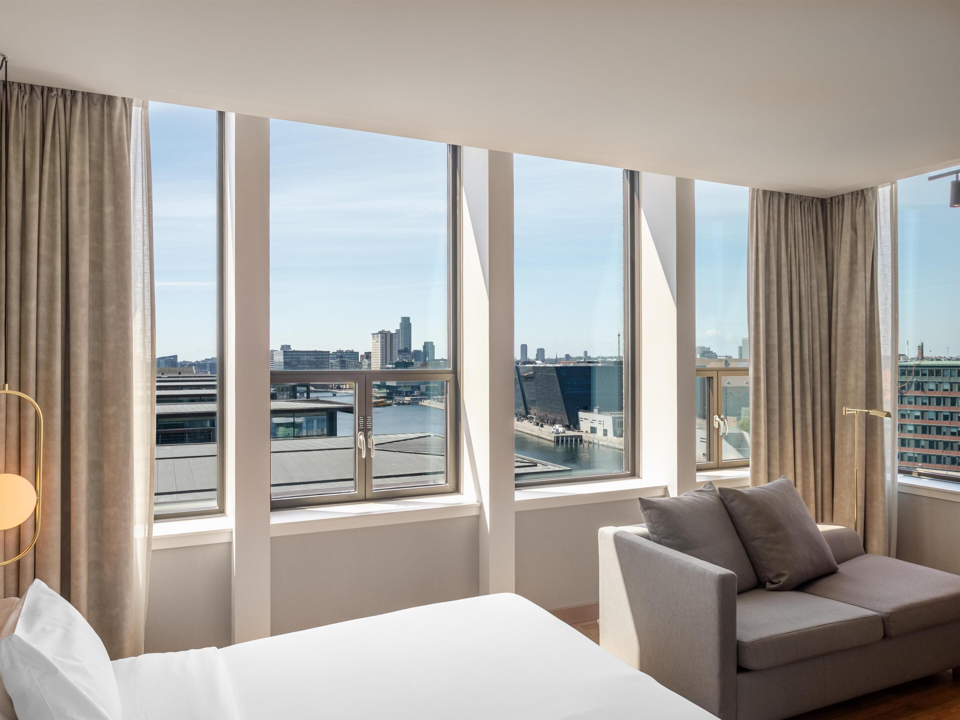A hotel room with a large window, a sofa, a bed, and a lamp.
