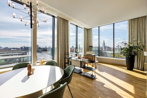 Habitación de hotel moderna con un ventanal que ofrece vistas de la ciudad, una mesa de comedor redonda y una estantería.