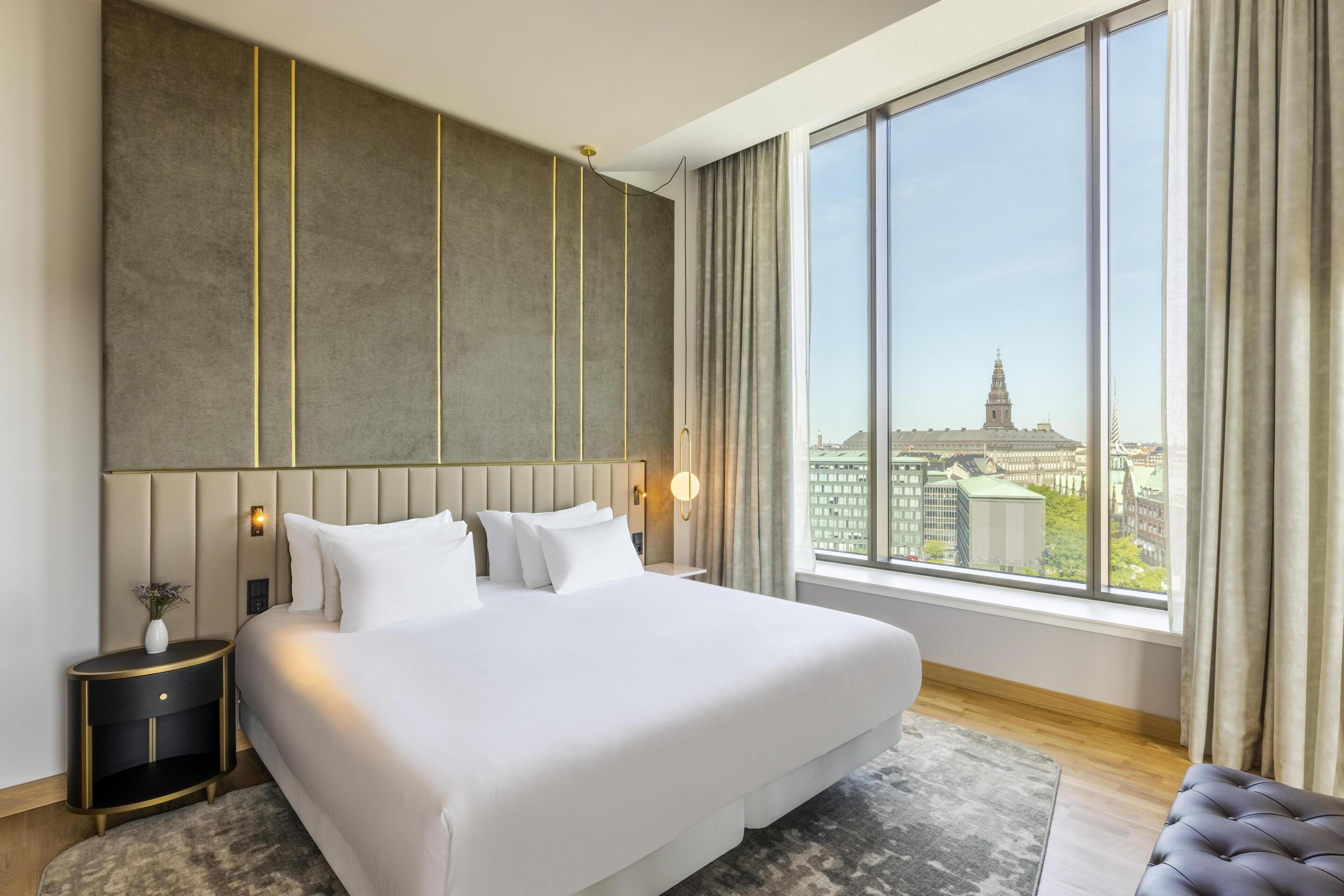 Une chambre d’hôtel moderne avec un grand lit, une table de chevet, une vue sur le paysage urbain à travers de grandes fenêtres et, au loin, une tour de la ville.