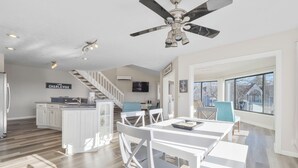 Une cuisine spacieuse dotée d’un îlot central, un coin repas avec des chaises blanches, un ventilateur de plafond et un escalier menant à l’étage supérieur.