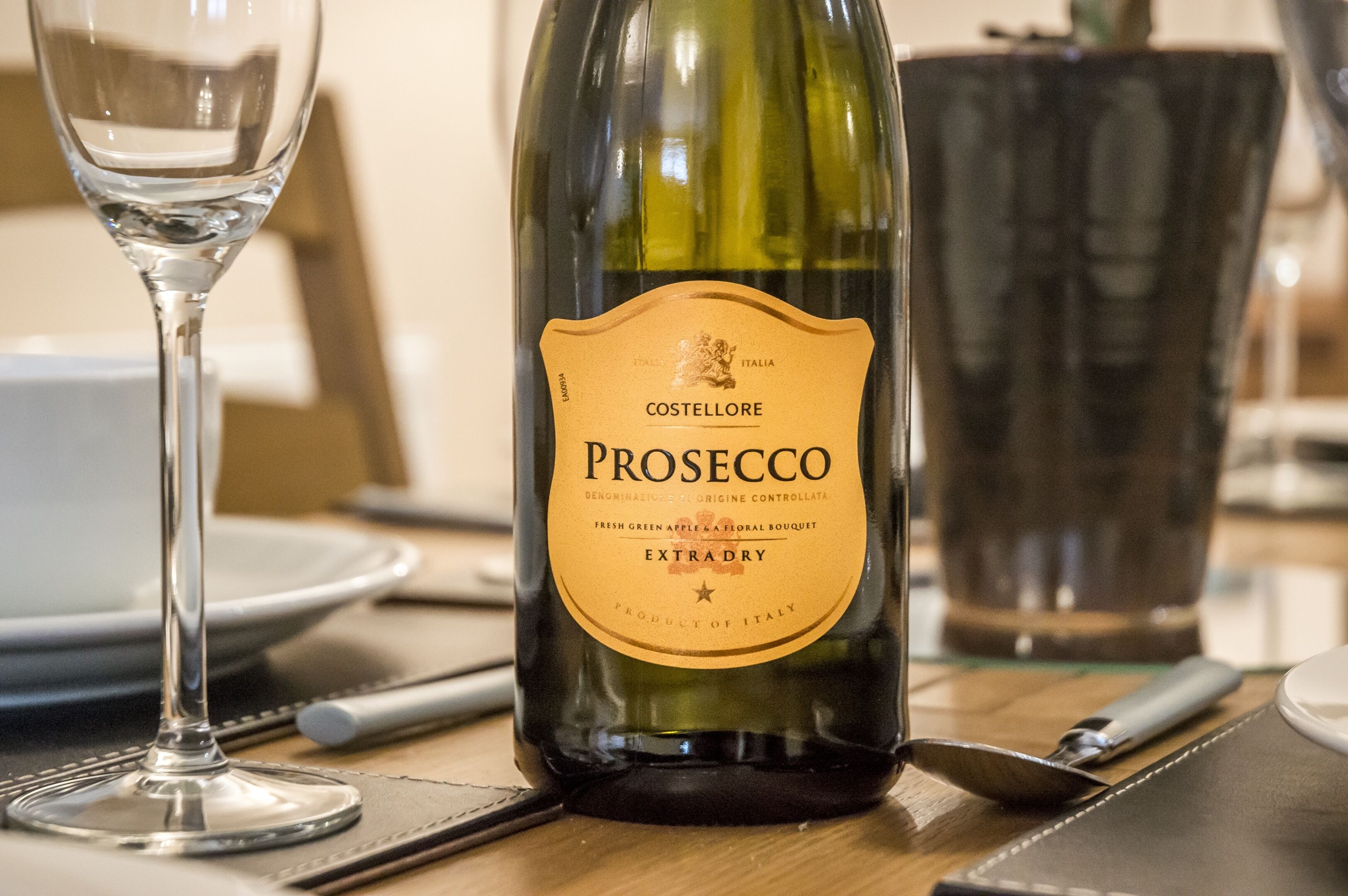 A bottle of Prosecco wine on a table with a glass and cutlery.