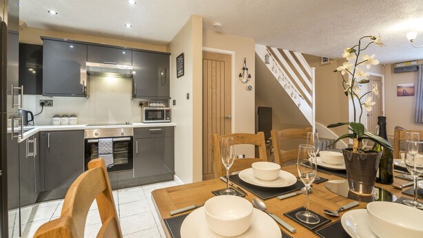 A modern kitchen with dark cabinets, a microwave, and a stove. There is a dining area with a wooden table set for two, including plates, glasses, and cutlery.