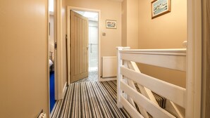 A hotel hallway with a striped carpet, a white railing, and a door leading to a room with a bed and a window.