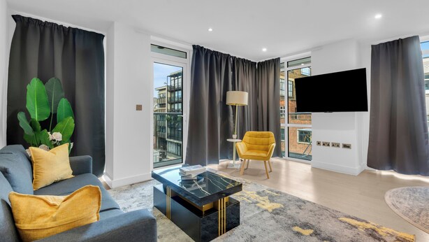 A modern living room with a sofa, coffee table, and armchair, large windows, and a flat-screen TV. - Two Bed Serviced Apt in Old Street (London)