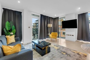 Una sala de estar moderna con sofá, mesa de centro y sillón, ventanas amplias y un televisor de pantalla plana.