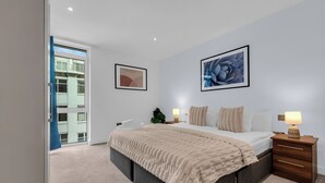 A modern bedroom with a large bed, a nightstand, and two wall-mounted lamps. - Two Bed Serviced Apt in Old Street (London)