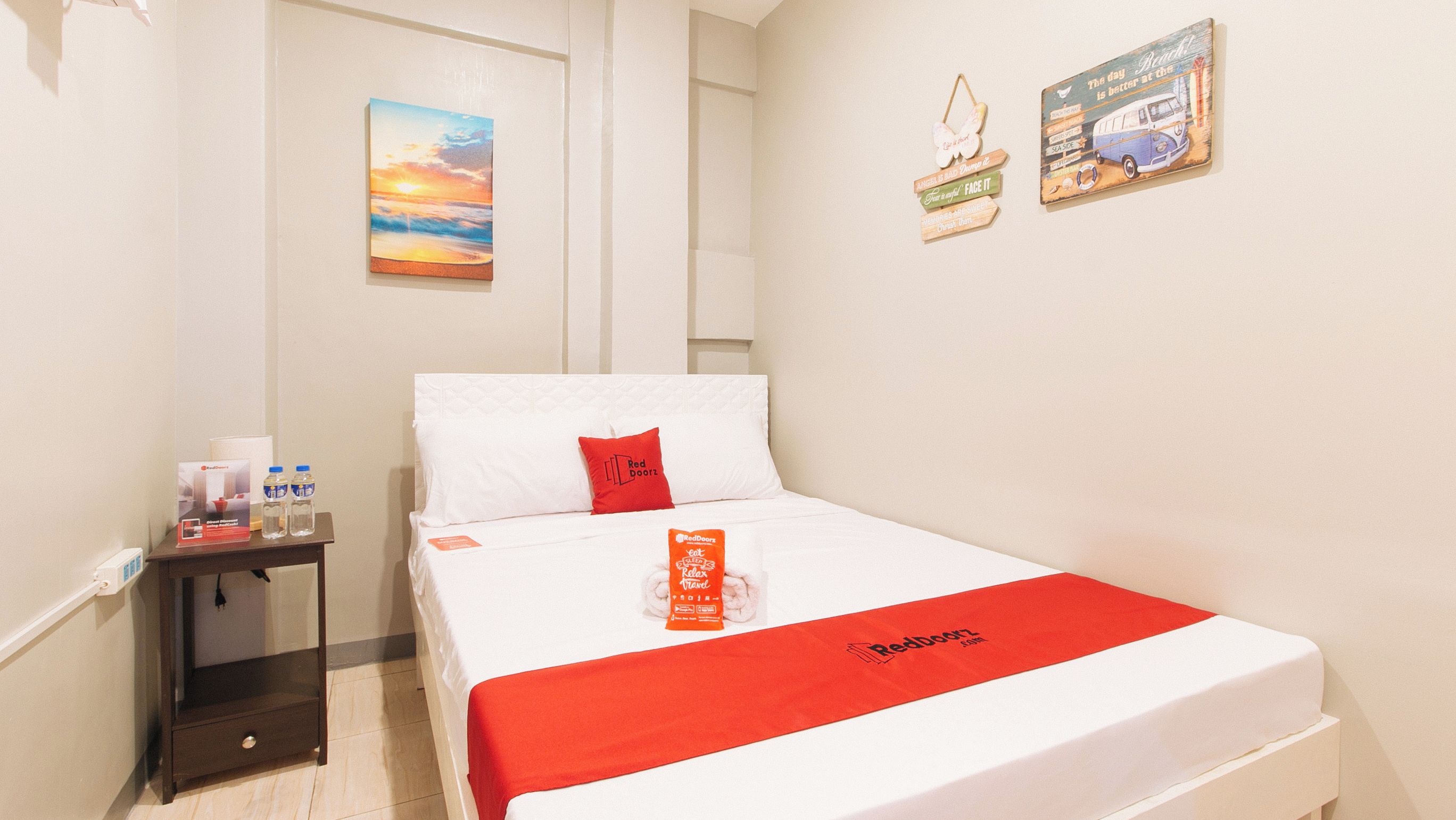 A hotel room with a bed, a nightstand, a water bottle, a RedDoorz sleep mask, and a wall with a sunset painting and two RedDoorz signs.