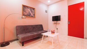 Una habitación con un sofá gris, una mesa blanca con botellas de agua, un cuadro en la pared, un televisor de pantalla plana y una puerta roja.