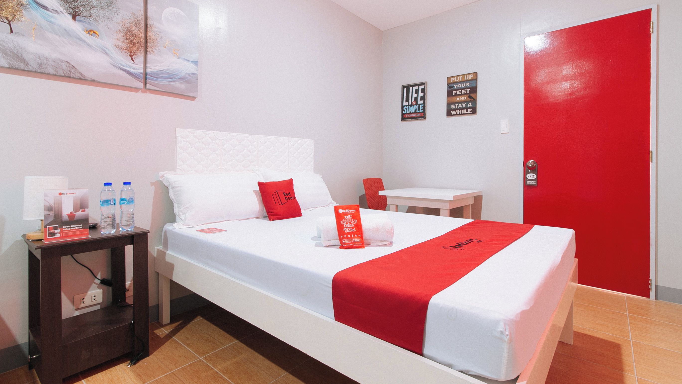 Une chambre d’hôtel avec un lit, une table de chevet avec des bouteilles d’eau, une porte rouge et un mur orné d’affiches motivantes.
