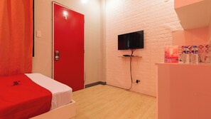 Une chambre d’hôtel avec une porte rouge, un lit recouvert d’une couverture blanche et rouge, un téléviseur à écran plat fixé au mur et une petite étagère avec des bouteilles d’eau.