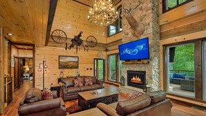 A spacious living room with a stone fireplace, leather sofas, and a large TV mounted on the wall. - Luxury Pool Cabin w Theater | Firepit | Hot tub (Sevierville)