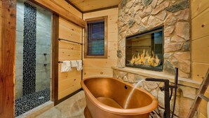 A wooden cabin interior with a stone wall, a fireplace, and a bathtub. - Luxury Pool Cabin w Theater | Firepit | Hot tub (Sevierville)