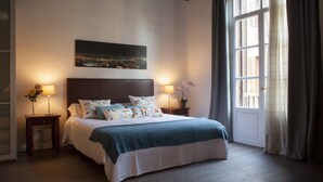 A bedroom with a large bed, two bedside tables with lamps, a cityscape painting on the wall, and a window with curtains. - Apartment Colom 1- (Barcelona)