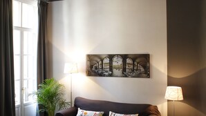 A living room with a dark brown sofa, a potted plant, a table, and a wall-mounted painting. - Apartment Colom 1- (Barcelona)