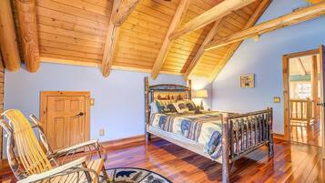 A bedroom with a sloped ceiling, wooden beams, a bed with a patterned bedspread, a wooden chair, and a door.
