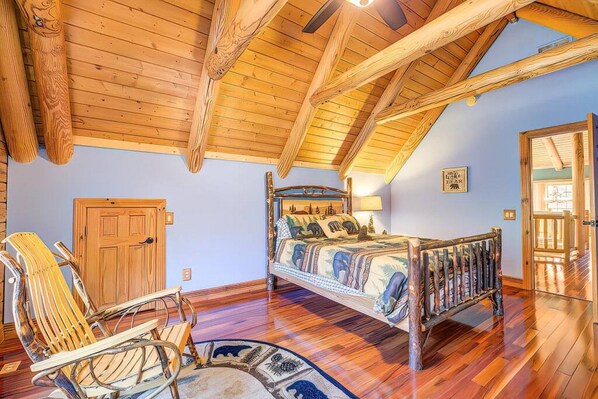 Une chambre avec un plafond en pente, des poutres en bois, un lit recouvert d’une couvre-lit à motifs, une chaise en bois et une porte.