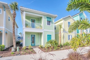 A two-story residential building with a balcony, a front door, and a small garden area with plants and a palm tree.