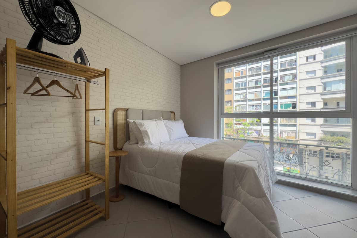Une chambre à coucher avec un lit, une étagère en bois, un ventilateur et une grande fenêtre donnant sur des immeubles.