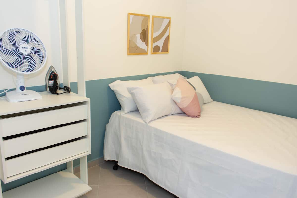 Une chambre à coucher avec un lit, une table de chevet sur laquelle se trouvent un ventilateur et un fer à repasser, et deux tableaux encadrés accrochés au mur.