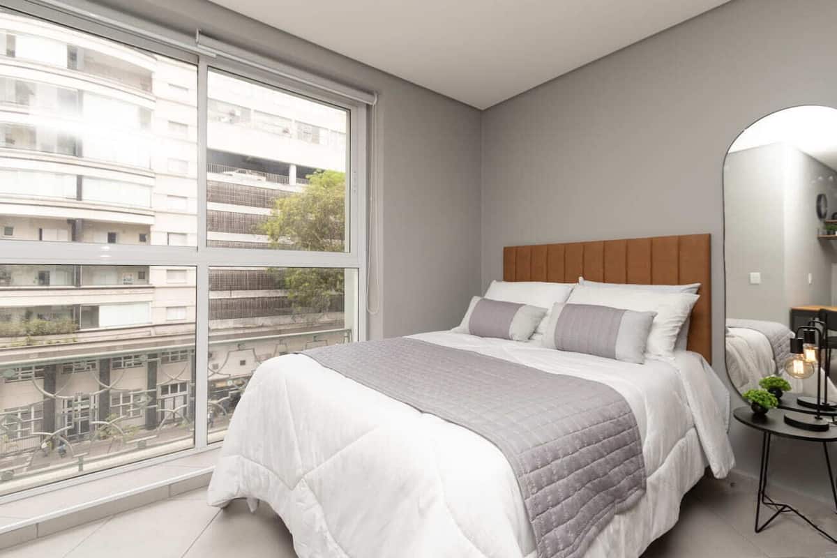 Une chambre moderne avec une grande fenêtre, un lit doté d’une tête de lit en bois et une table de chevet avec une lampe.