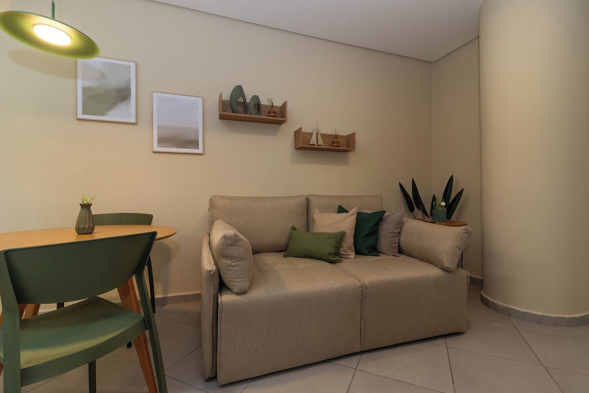 Un salon avec un canapé beige, une table basse en bois et un fauteuil vert. On y trouve deux étagères murales ornées d’objets décoratifs et deux tableaux encadrés.