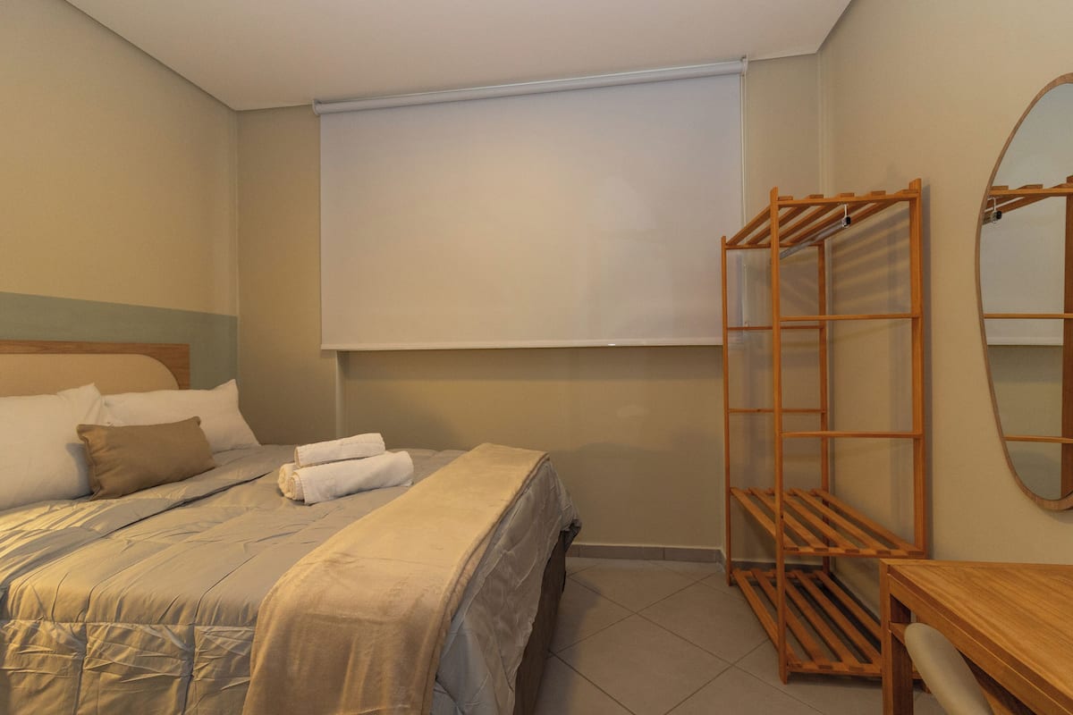 Une chambre d’hôtel avec un lit, un étendoir en bois, un miroir et une fenêtre avec un store.