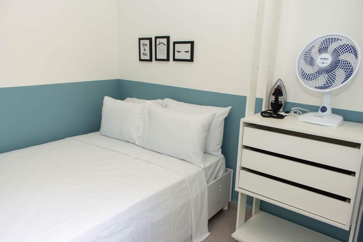 Un lit bien fait, avec du linge de lit blanc, une table de chevet blanche sur laquelle se trouvent un fer à repasser et un ventilateur, et trois tableaux encadrés accrochés au mur.