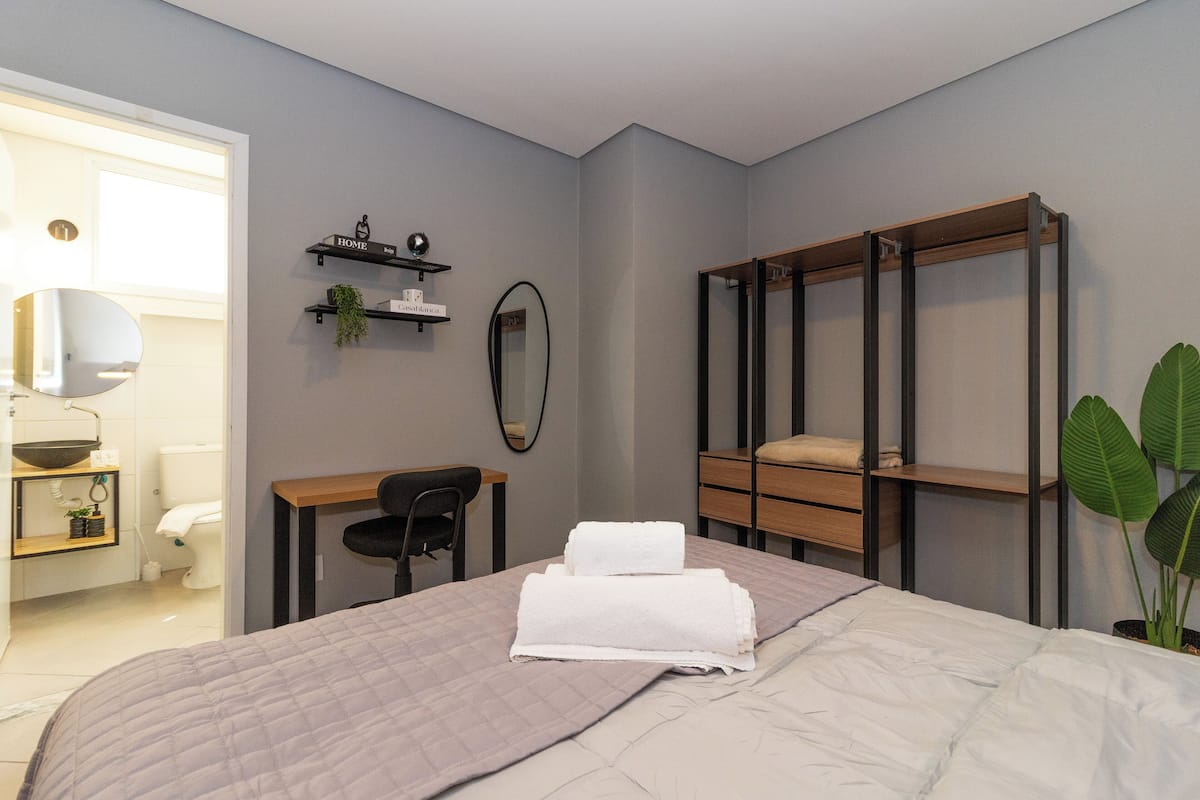 Une chambre à coucher moderne, avec un lit, un bureau, une chaise, une armoire et une salle de bain visible à travers une porte ouverte.