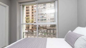 A bedroom with a large window, a bed with a gray blanket, and a view of a building with balconies. - 360 Av. Ipiranga (São Paulo)
