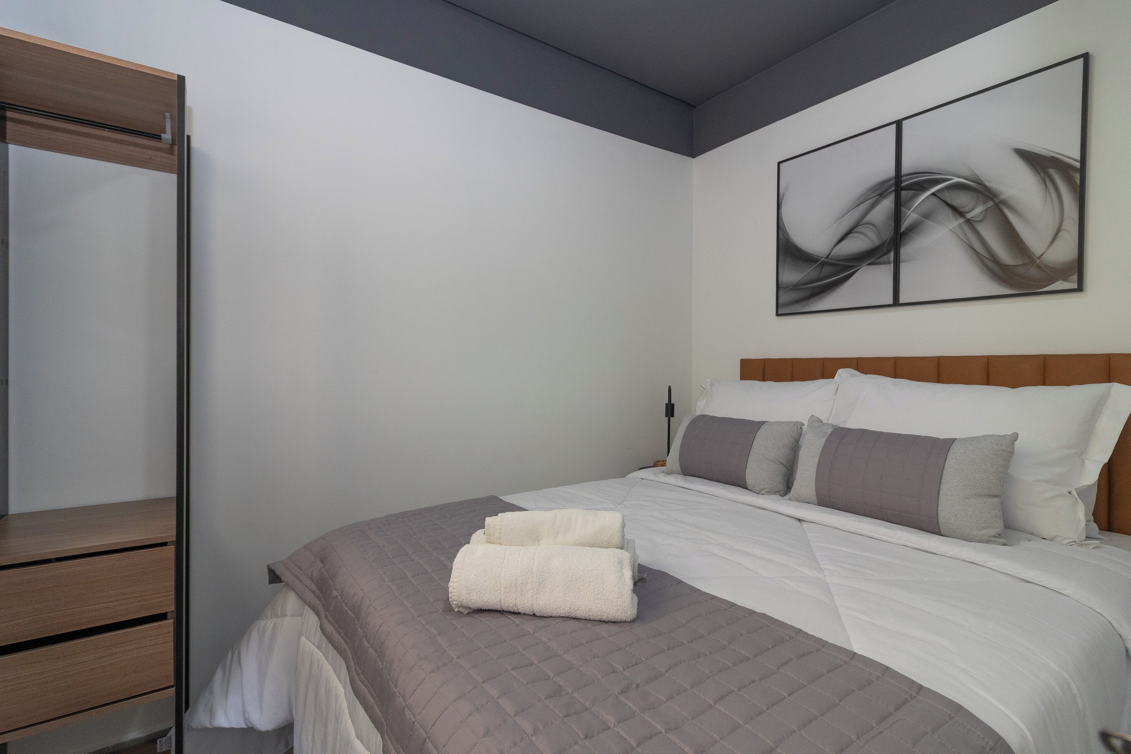 A neatly made bed with a grey quilt, a white pillow, and a grey pillow on a wooden headboard. A white towel is folded on the bed. A large abstract painting is on the wall above the bed.
