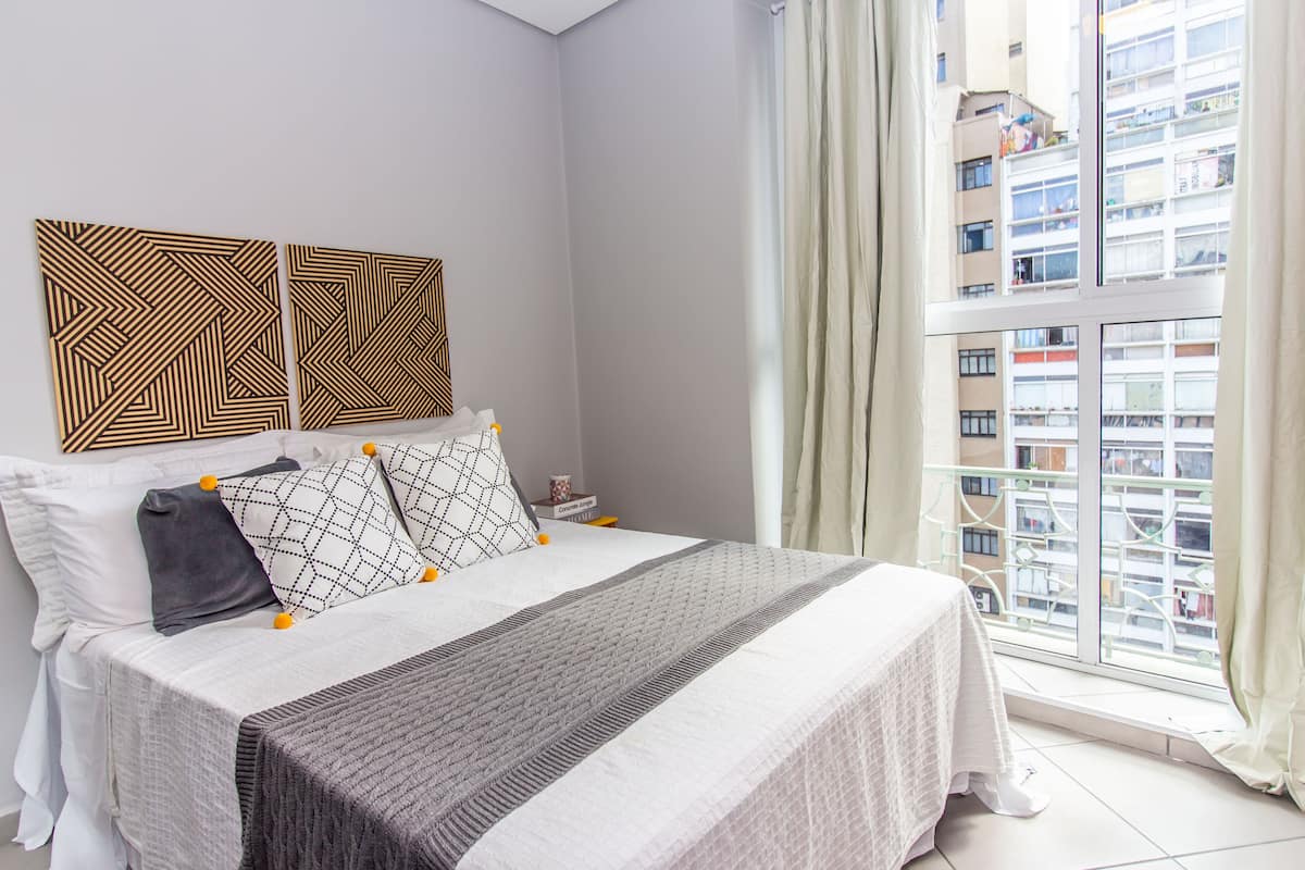 Un lit bien fait, avec une tête de lit à motifs, une table de chevet avec une lampe, et une vue sur un immeuble de grande hauteur par la fenêtre.