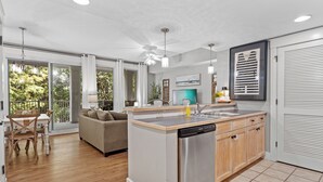 A modern kitchen with a dishwasher, wooden cabinets, and a dining area with a table and chairs. - 1BR Condo at Baytowne! Steps to Wharf, Tropical Pool & Hot Tub, Balcony View. (Miramar Beach)