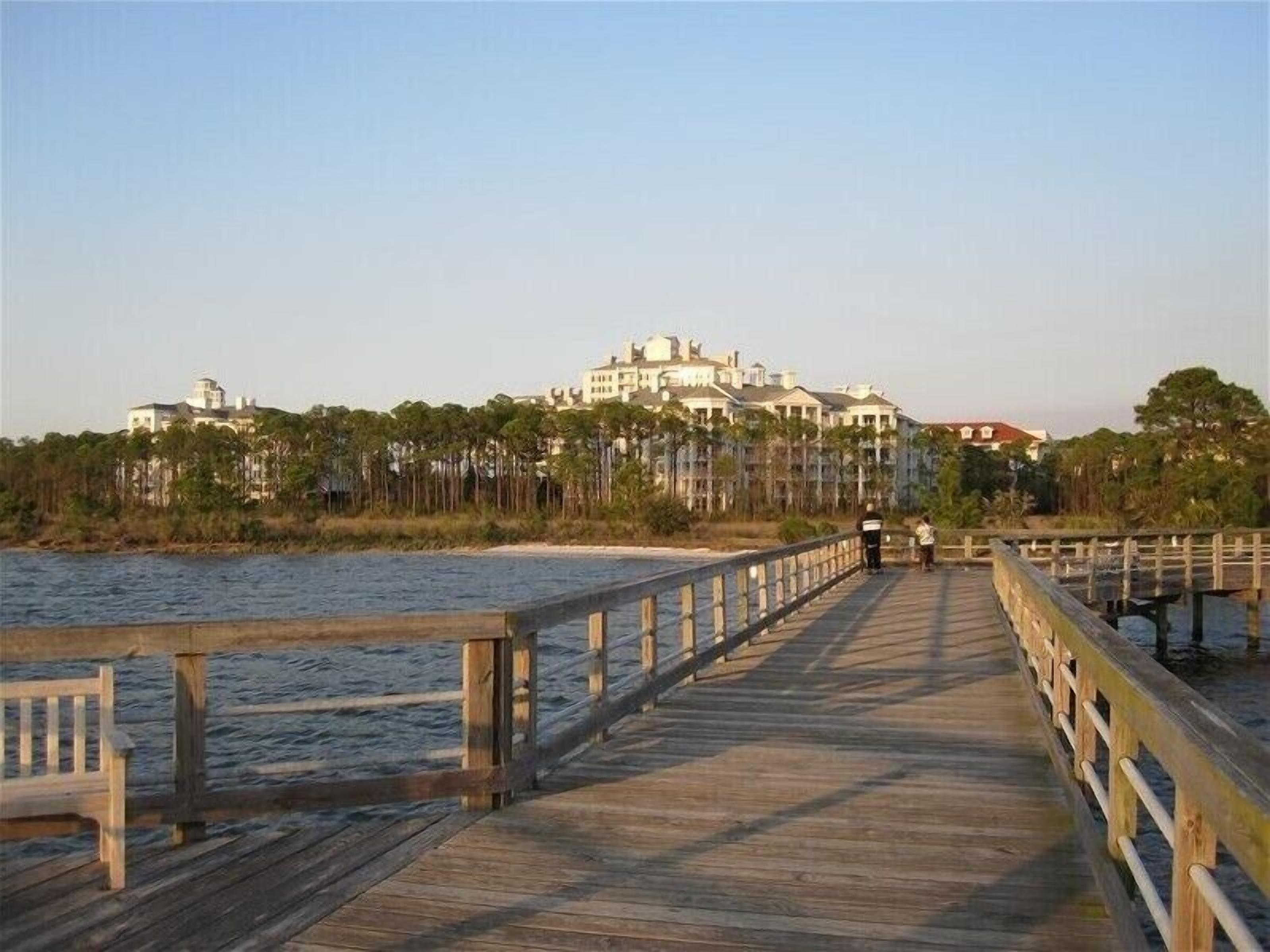 A wooden pier leading to a beachfront property with multiple buildings, a sandy beach, and a forested area.