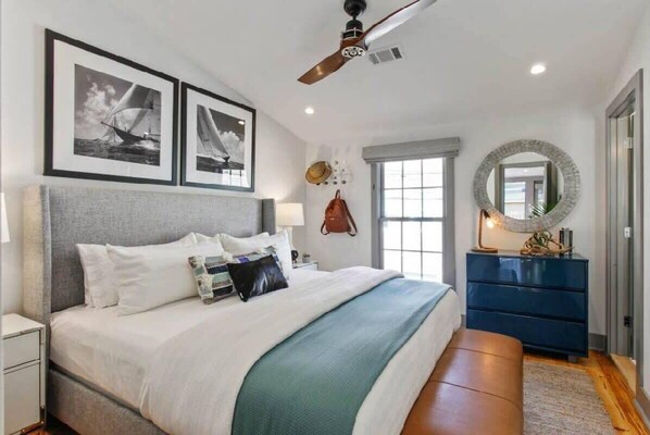 A bedroom with a large bed, a ceiling fan, framed pictures on the wall, a window, and a dresser.