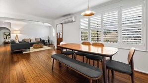 A modern living room with a dining area, wooden flooring, and large windows with blinds. - Bendalong | Rural Retreat, Short Drive to CBD (Nashdale)