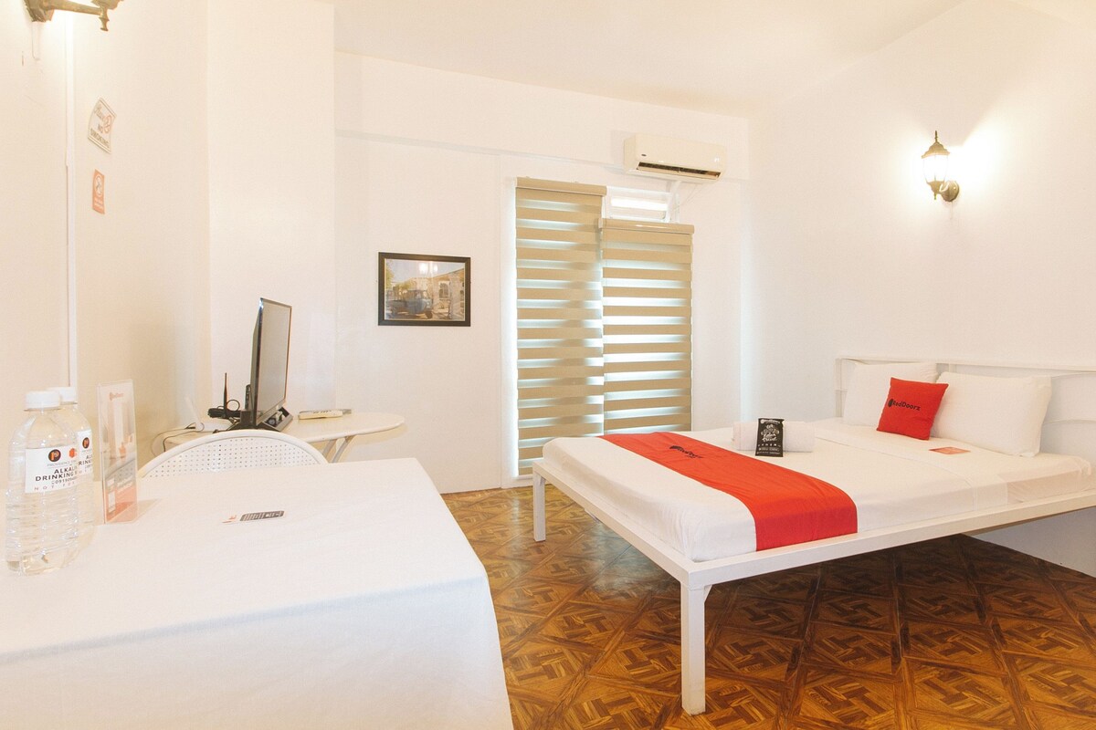 A hotel room with a bed, a desk with a chair, a table with water bottles, a picture on the wall, and a window with blinds.