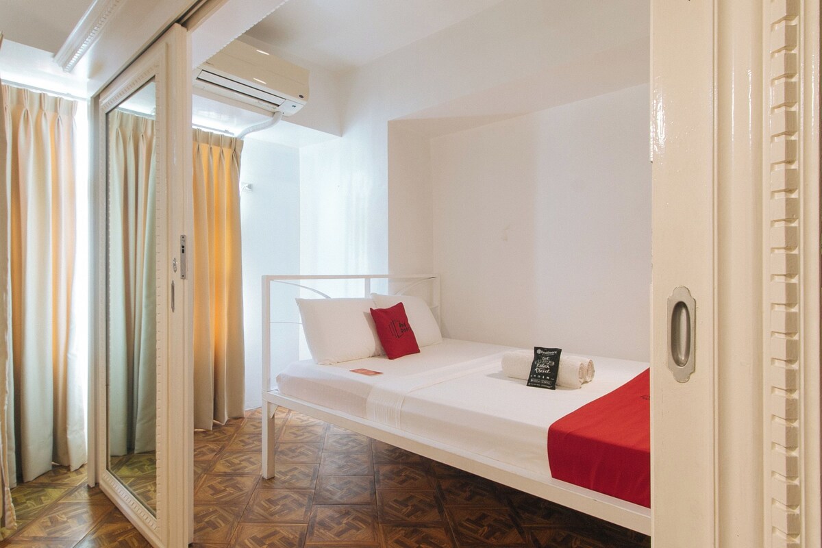 A single bed with a red pillow and a towel, a white headboard, and a sliding glass door leading to a balcony.