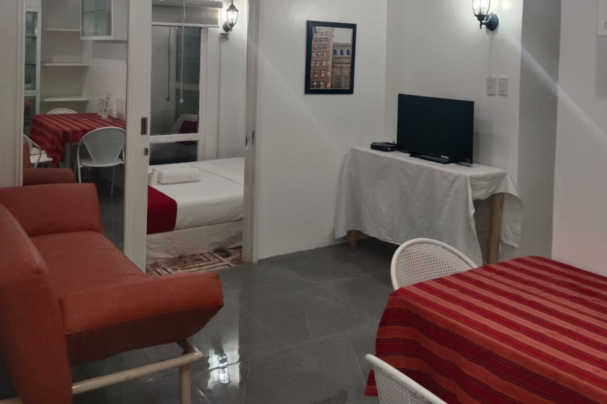 A hotel room with a bed, a TV on a small table, a sofa, and a dining area with a table and chairs.