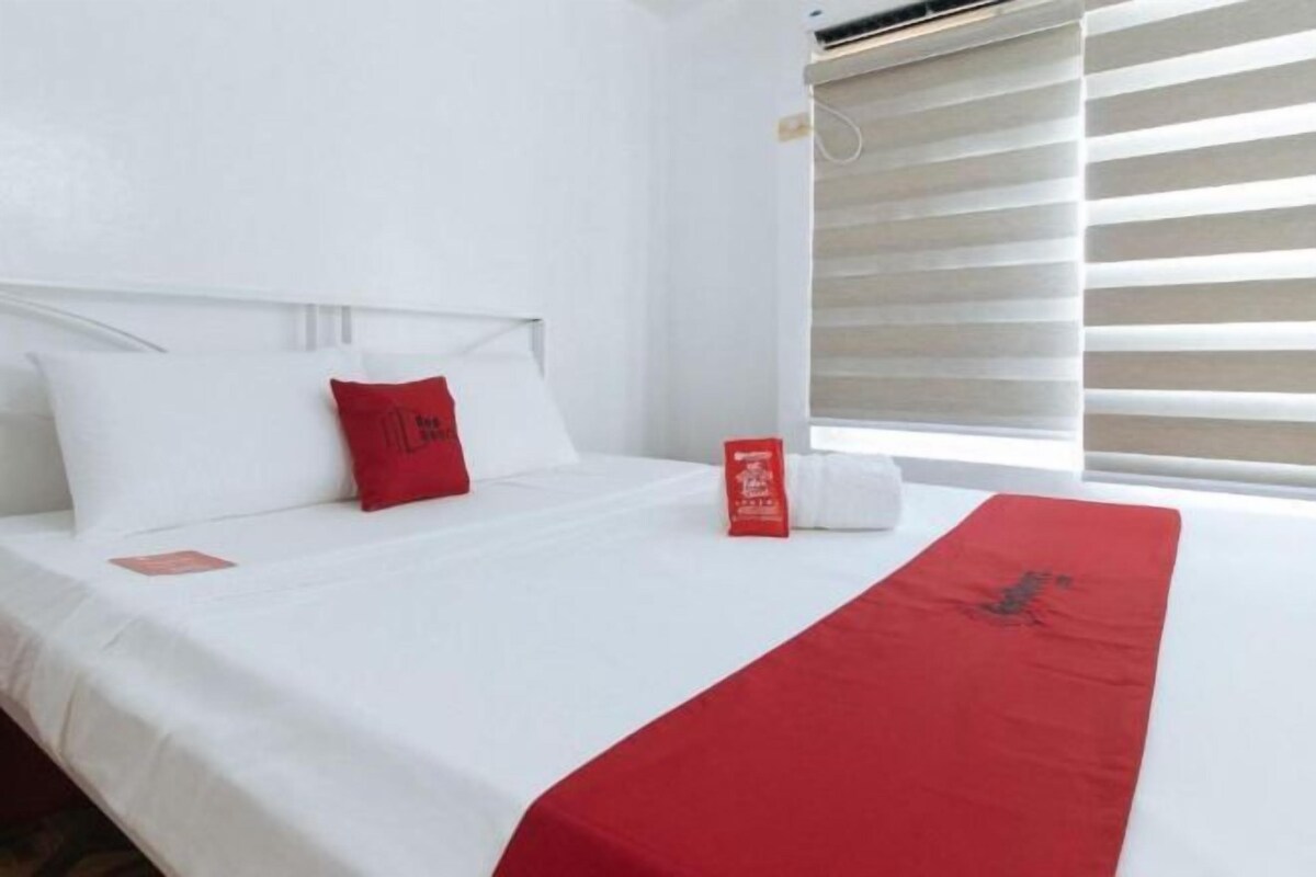 A hotel room with a neatly made bed, a red pillow, a white pillow, and a red throw blanket.