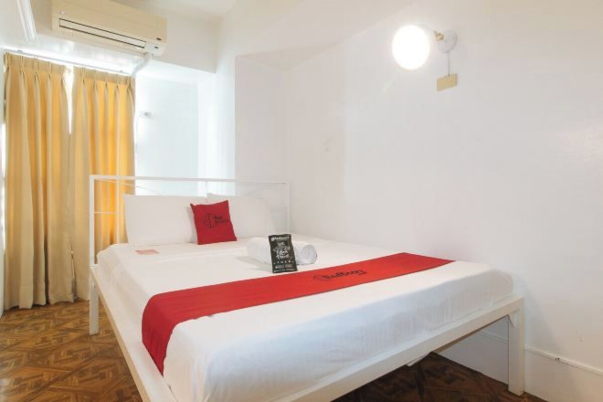 A hotel room with a bed, a red throw, a pillow, a bedside table, and a wall-mounted light.
