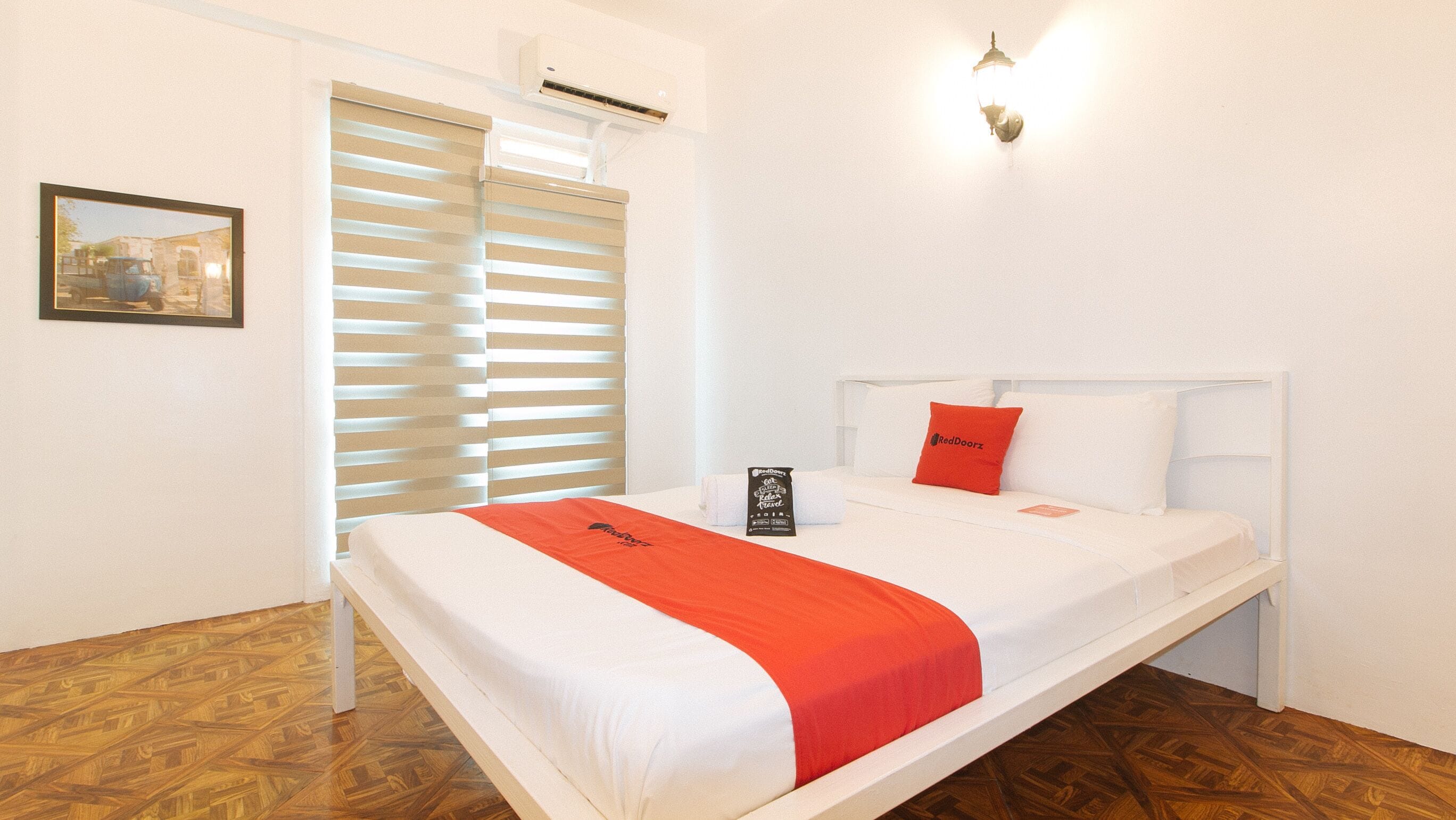 A hotel room with a bed, a window with blinds, an air conditioning unit, and a wall-mounted light fixture.