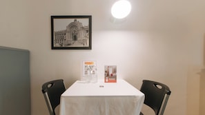 A table with a white tablecloth, two black chairs, a framed black and white photo of a building, and two RedDoorz promotional stands. - Reddoorz @ The Providence Tower (Manila)