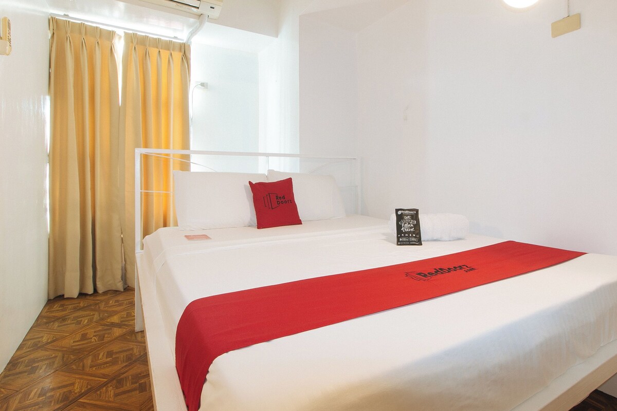 A hotel room with a bed, a red throw, a pillow with a red logo, a white wall, and a window with curtains.