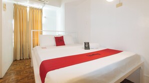 A hotel room with a bed, a red throw, a pillow with a red logo, a white wall, and a window with curtains. - Reddoorz @ The Providence Tower (Manila)