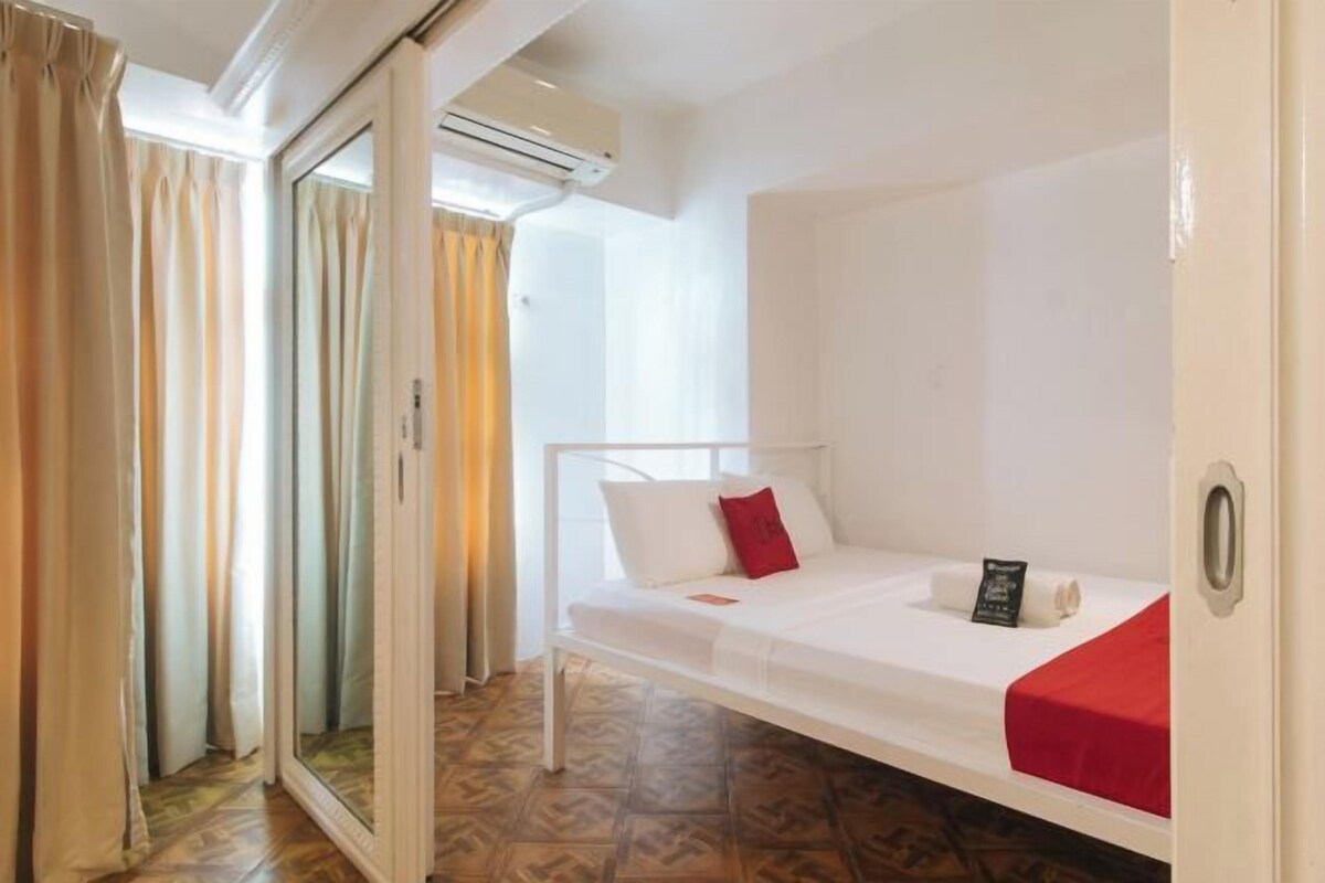 A single-bed room with a white bed, red pillow, and a small bedside table with a book.