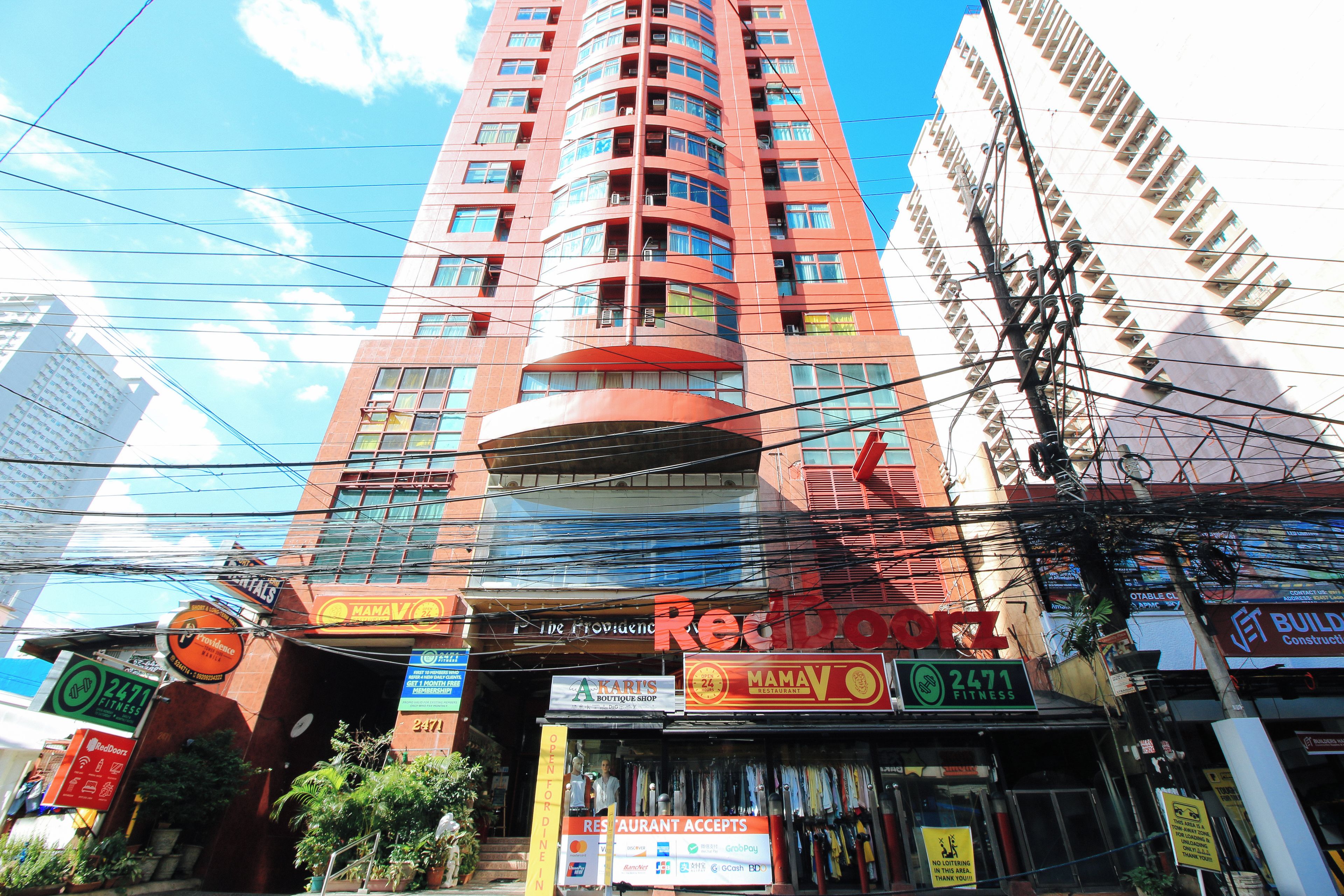 Um edifício com vários andares, fachada vermelha marcante, diversas varandas e variedade de estabelecimentos comerciais no andar térreo, incluindo um centro de fitness e um restaurante.