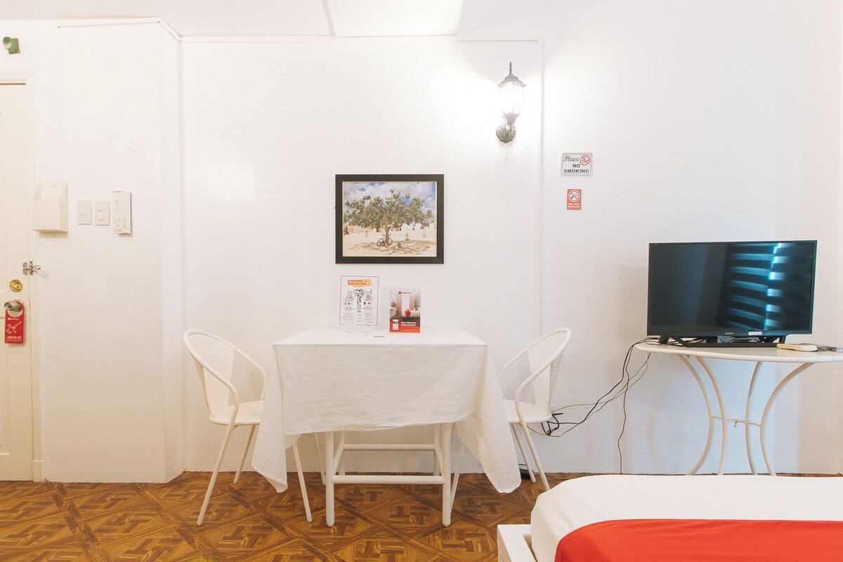 A hotel room with a bed, a TV, a table with chairs, a framed picture on the wall, and a door with a red sign.