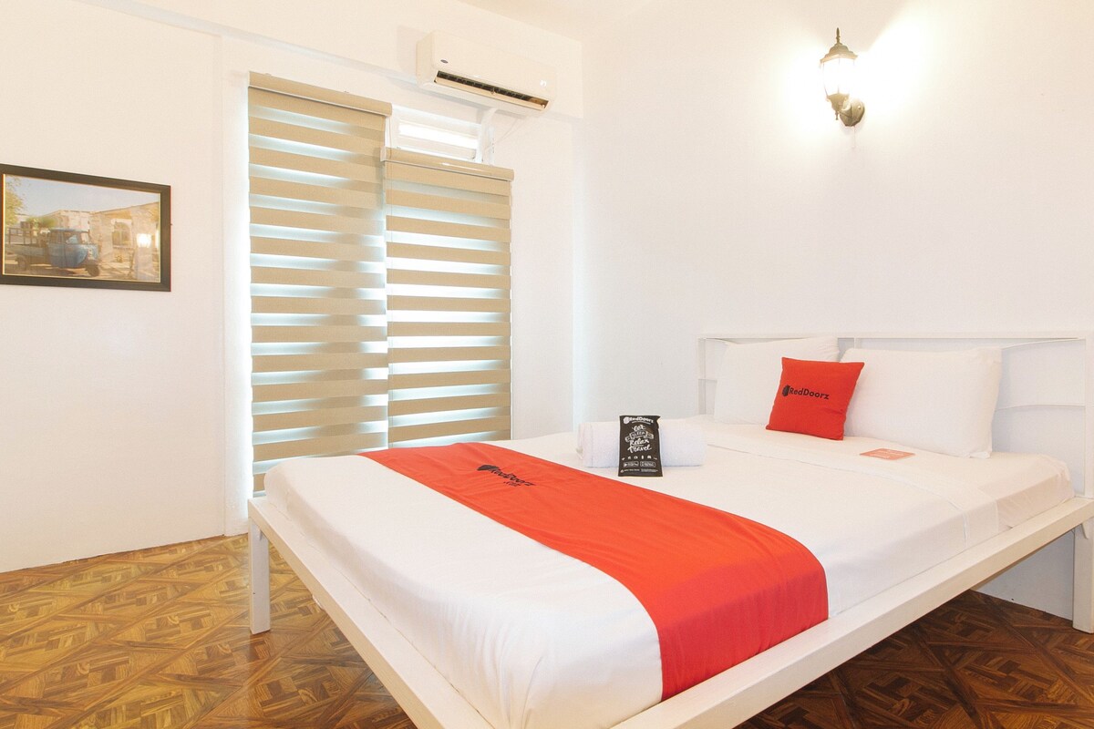 Quarto de hotel com cama, janela com persianas, ar condicionado, luminária fixada na parede e quadro emoldurado.