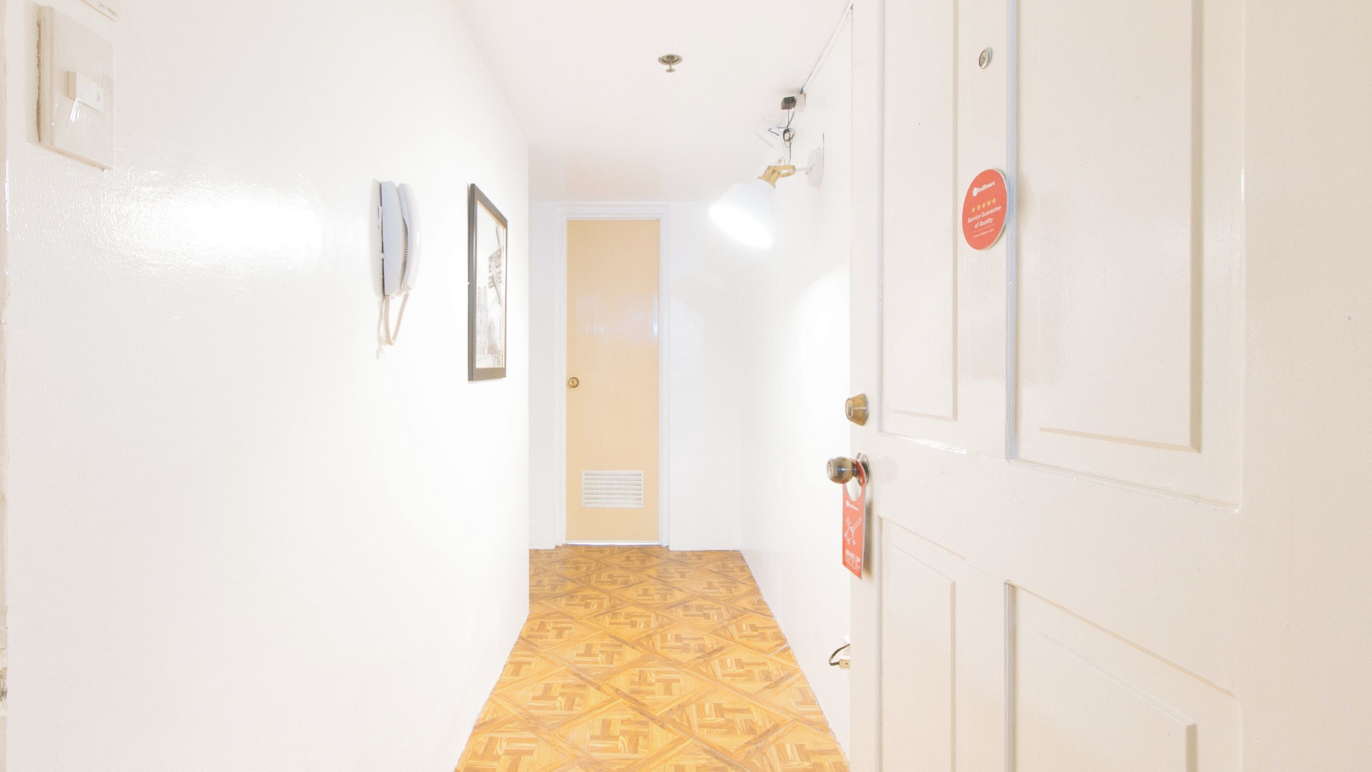 Un pasillo con piso estampado, paredes blancas, una puerta con un letrero y una lámpara.
