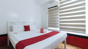 A hotel room with a bed, a red pillow, a white pillow, a red throw blanket, a white pillowcase, a white bedside table, and a window with blinds. - Reddoorz @ The Providence Tower (Manila)